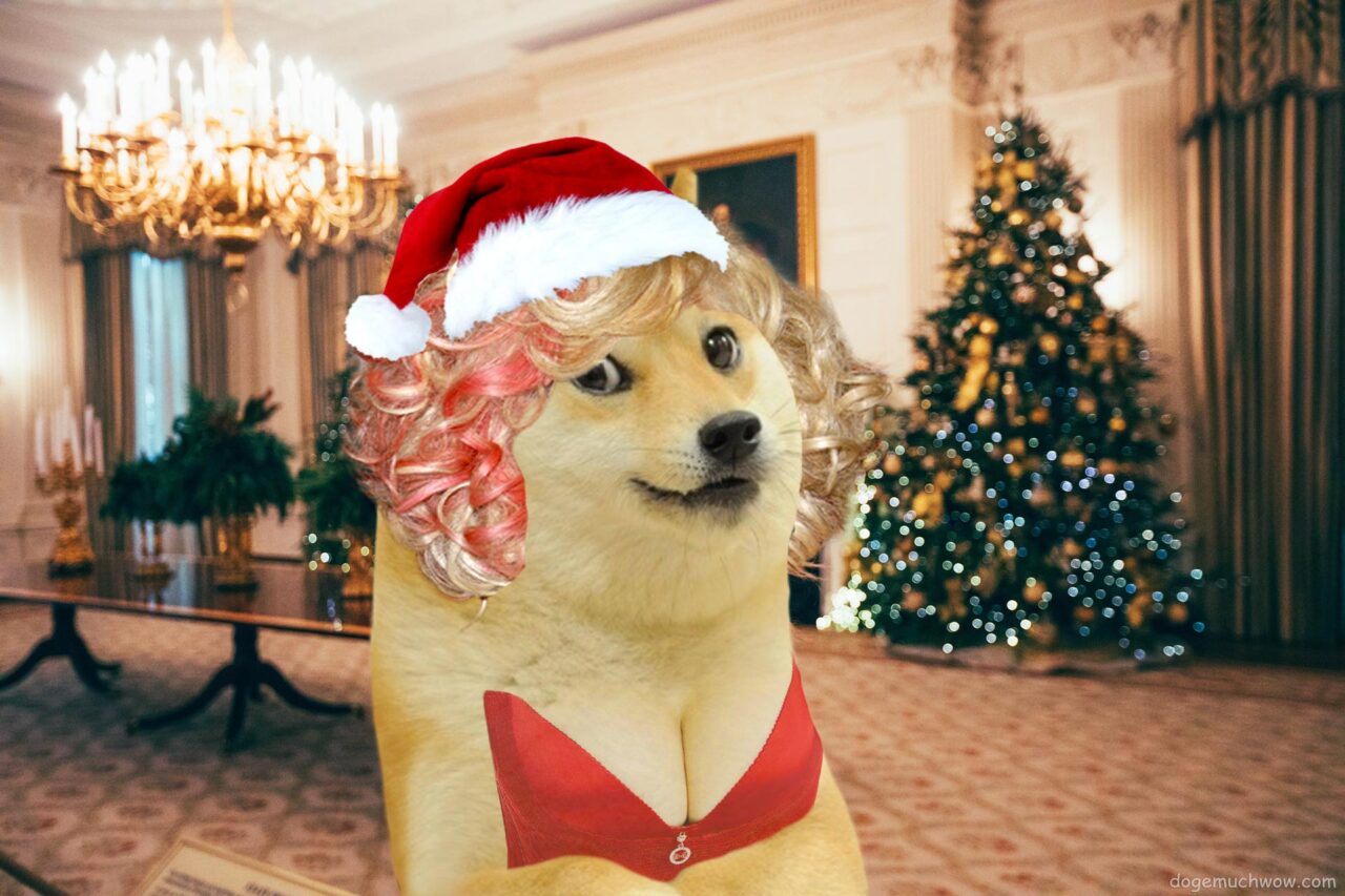 Mrs Claus with blonde-pink hair, red dress and santa's hat posing in her Christmas residence. In the background, a beautifully lit Christmas tree and a chandelier.